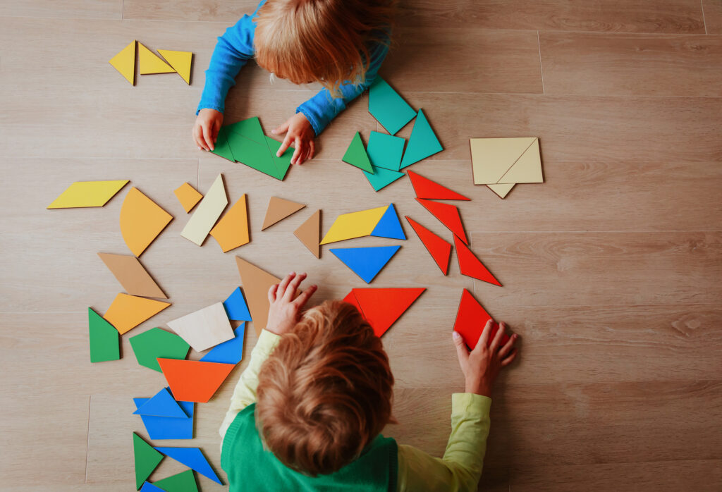Two children laying on the floor facing each other with a pile of mosaic shape tiles in between them
