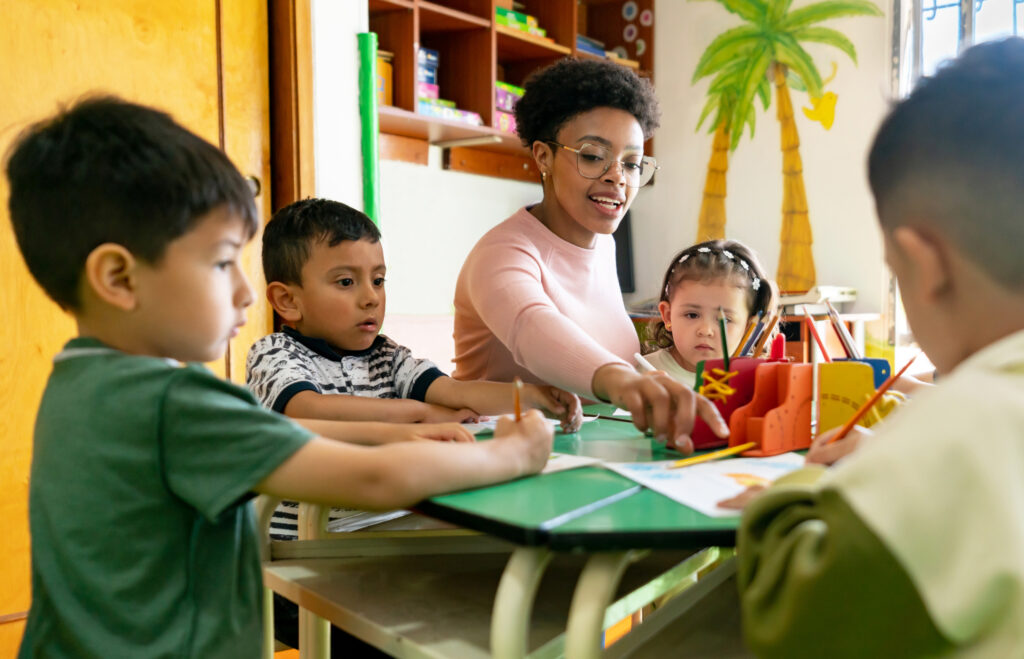 Teacher seated with group of toddlers, pointing at a coloring sheet