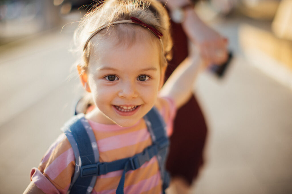 Close up of toddler girl wearing a backpack, reaching behind to hold her parent's hand